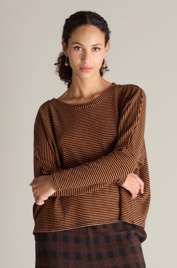 A person with curly hair wearing a Mini Stripe Boxy Top and a checkered skirt stands with folded arms against a plain background, showcasing a versatile wardrobe essential.