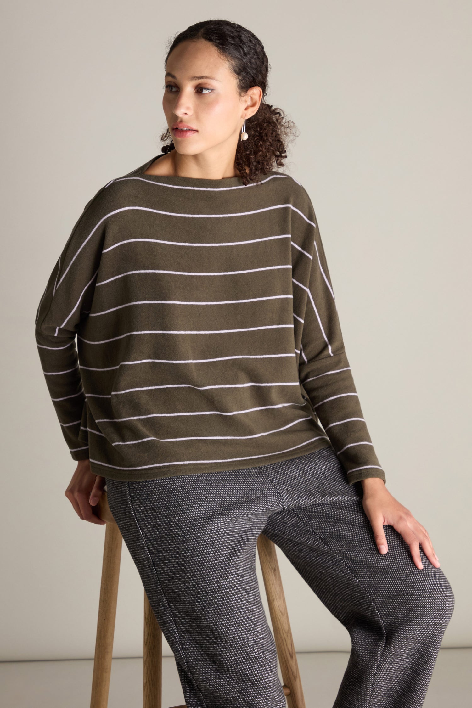 A person in a striped olive sweater and gray pants sits on a stool against a plain background, wearing a Neirami Narrow Stripe Boat Neck Boxy Top.