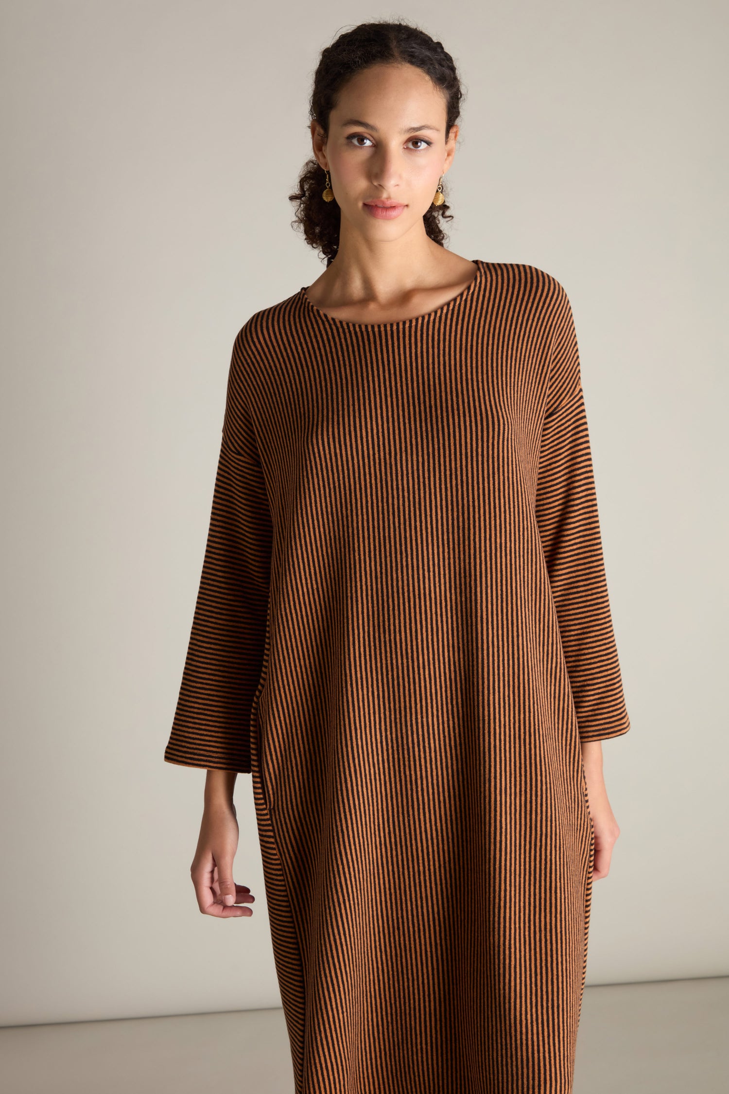 A person with dark curly hair is wearing the Mini Stripe Flared Dress in brown and black, featuring a relaxed drop shoulder design, standing against a plain background.