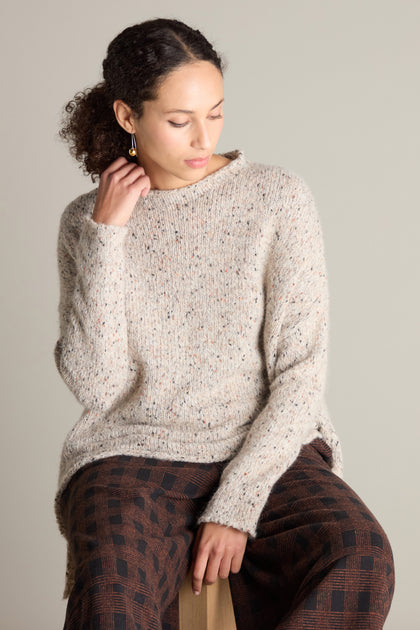 A person with curly hair is seated, wearing a Neirami knitted sweater and patterned pants, looking down with one hand touching the Marl Scoop Neck Knit.