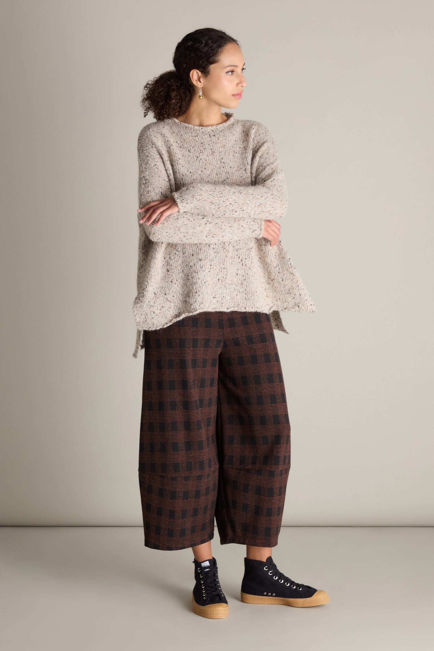 A person stands against a plain backdrop, wearing a beige Marl Scoop Neck Knit from Neirami, brown checkered culottes, and black sneakers with beige soles. They have their arms folded and are looking to the side.