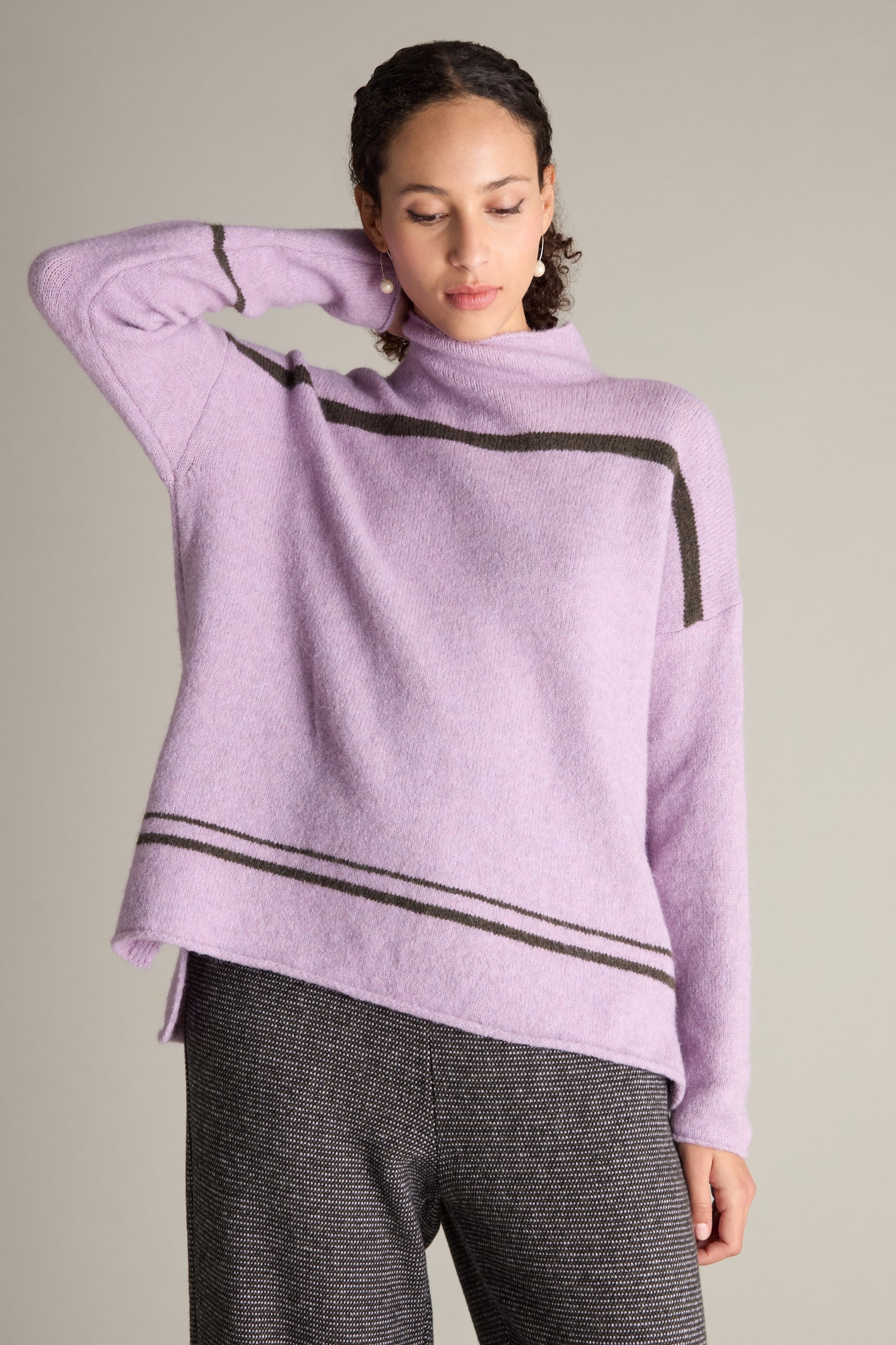 A person poses with one hand behind their head, wearing a contemporary style light purple Stripe Detail Knit sweater featuring black stripes and paired with gray pants against a plain background, exuding cosy comfort.