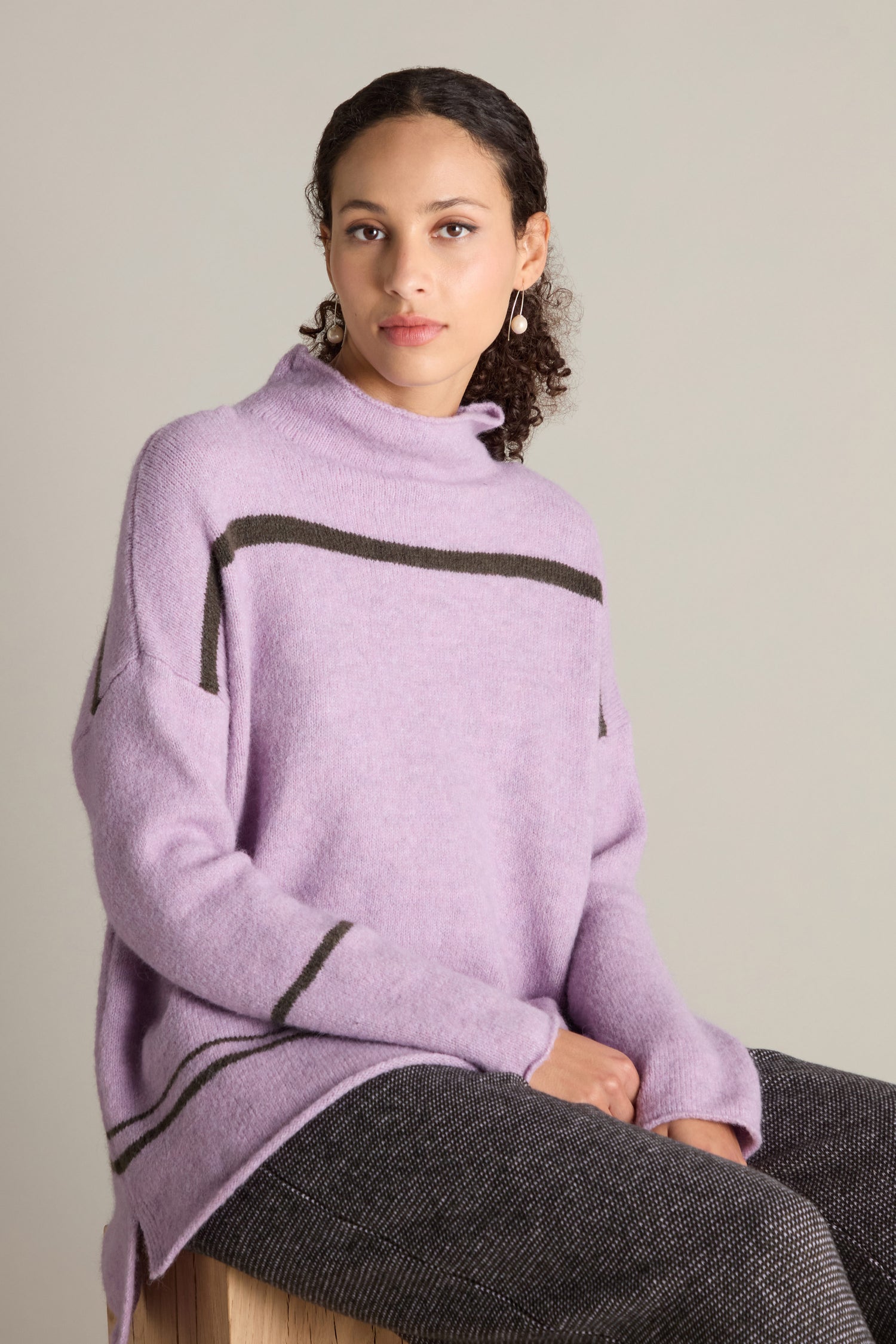 A woman with curly hair, dressed in a cozy lavender sweater called "Stripe Detail Knit" and gray pants, sits on a wooden stool against a neutral background.