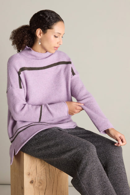 A person with curly hair sits on a wooden block, glancing down at their sleeve. Clad in the stylish Stripe Detail Knit sweater featuring black stripes and paired with gray pants, they embody cosy comfort in a contemporary look.