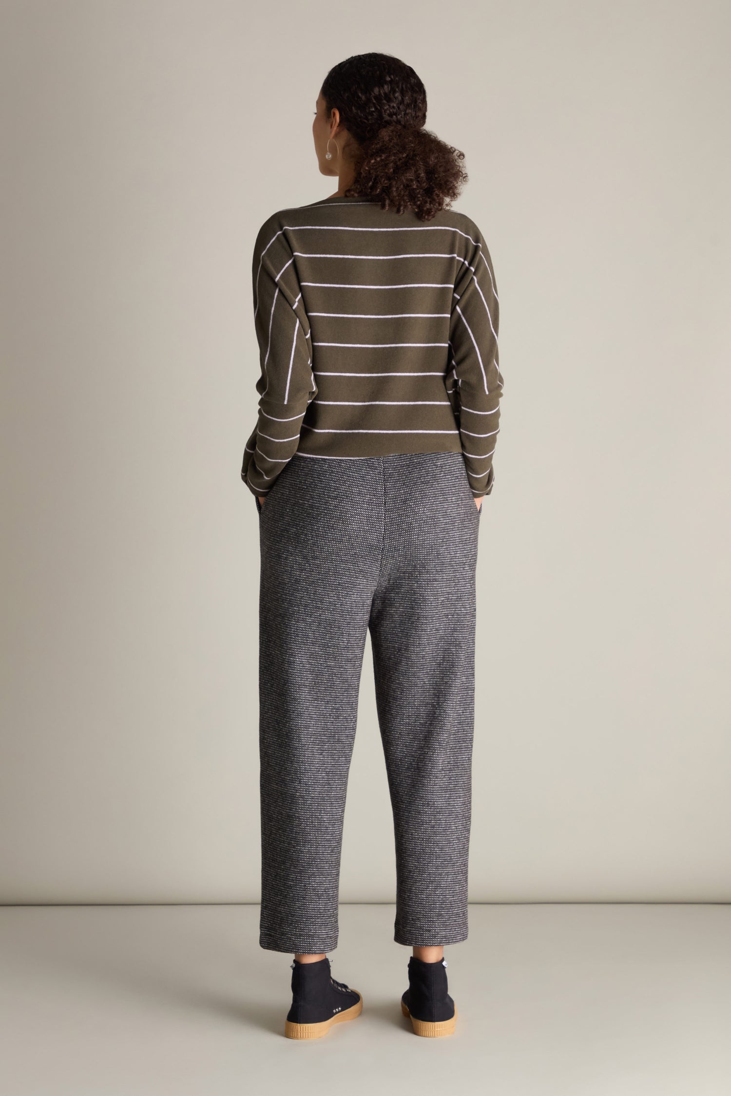 A person is standing in a minimalistic setting, seen from the back. They are wearing a green and white striped sweater, chic Jaquard Jersey Trousers featuring a cropped length and stretch waistband, along with black shoes. Their hands appear to be in their pockets.