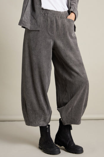 A person is wearing the Stretch Cord Bubble Trouser, featuring a loose-fitting design in gray corduroy with an elasticated waistband, paired with black boots. Their hands rest in the pockets of the trousers. The upper body and head are not visible.