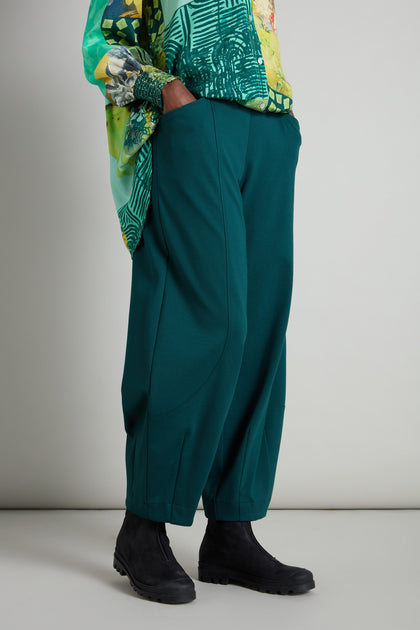 A person wears a patterned green top crafted from premium jersey fabric, paired with teal Jersey Bubble Trousers and black boots, set against a neutral backdrop with a touch of Danish-inspired style.