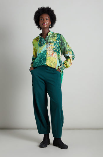 A person wearing a vibrant, patterned blouse and dark green Jersey Bubble Trousers stands against a plain background, hands in pockets, showcasing a Danish-inspired design flair.