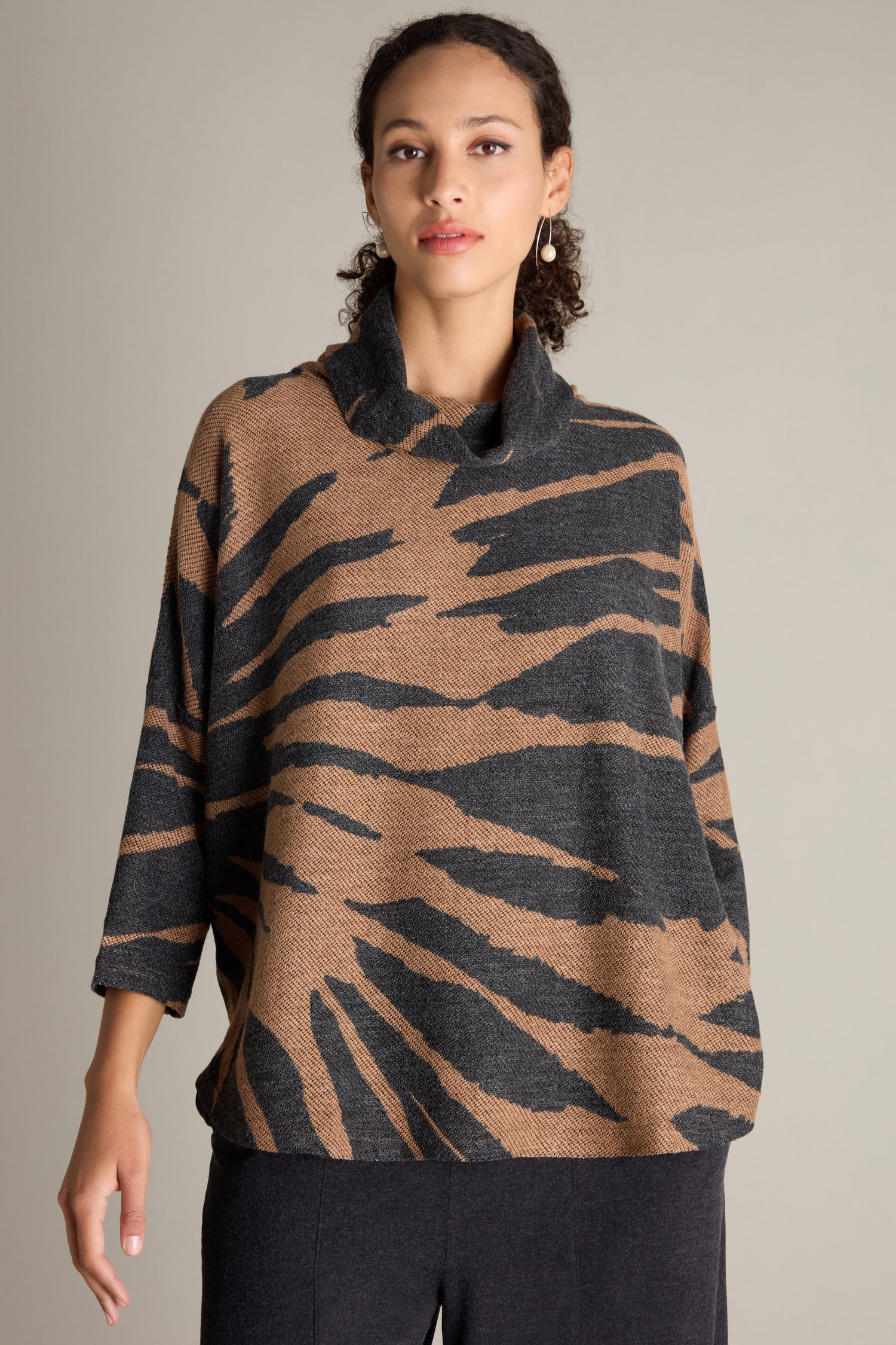 A person with curly hair is wearing the Cowl Neck Jacquard Top, which features a loose fit and long sleeves. The luxe jacquard fabric is adorned with an abstract black and brown striped pattern against a neutral background.