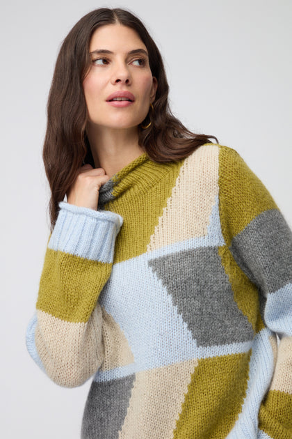 Woman with long brown hair wears the Blue Colour Block Knit sweater, featuring green, blue, beige, and gray panels, as she looks off to the side against a plain background.