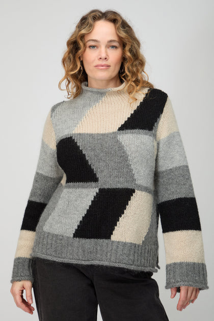 A woman models the Grey Colour Block Knit, a loose sweater in grey, black, and beige tones, embodying Scandinavian minimalism against a light background.