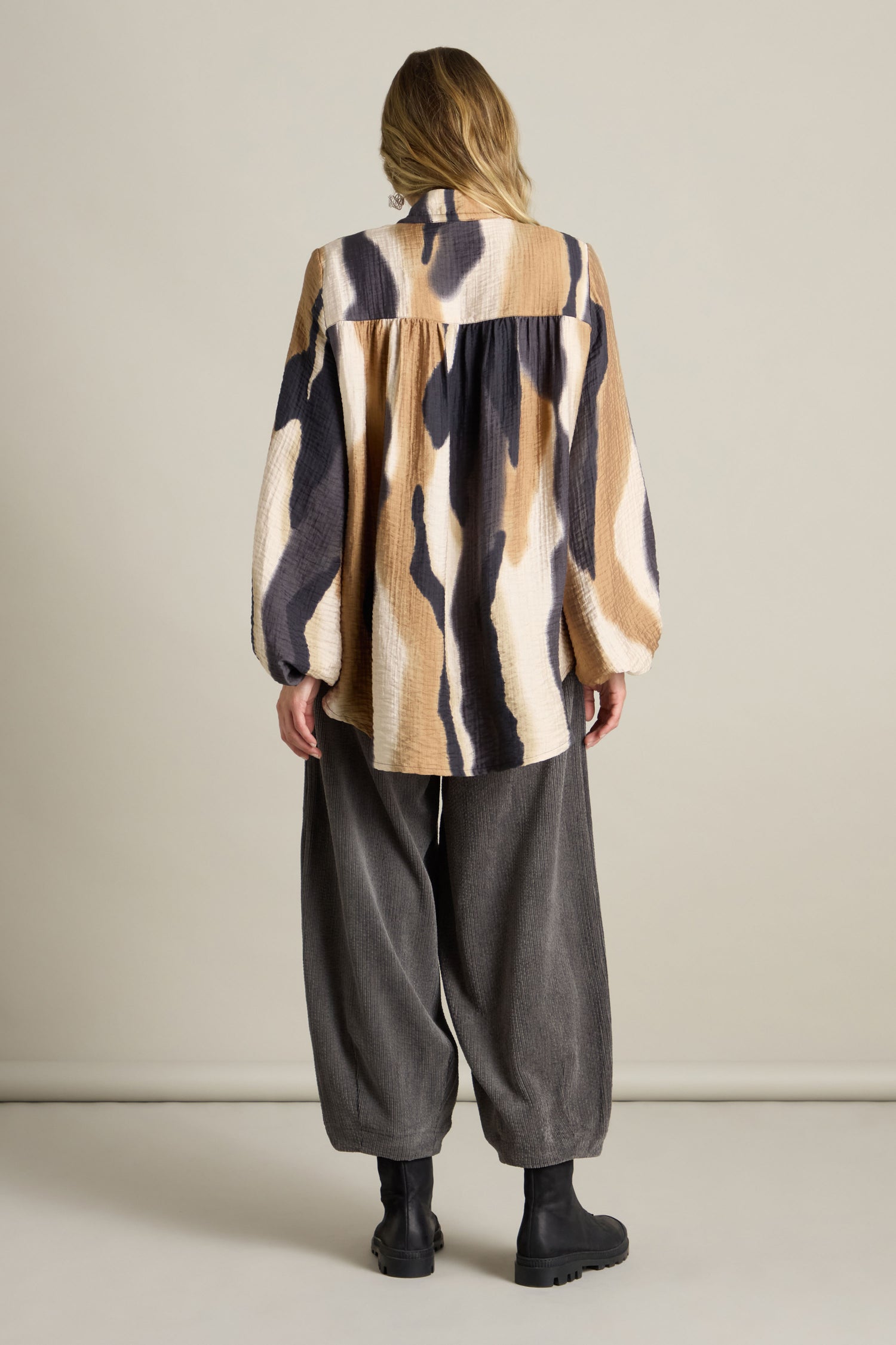 A person stands indoors facing away, wearing a beige and black patterned long-sleeve shirt, loose gray pants, and black boots. The Crinkle Viscose Print Shirt adds a textured feel to their ensemble.