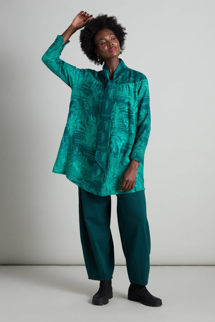 Wearing a relaxed Tie Neck Viscose Shirt in a lush green jungle print, paired with matching pants and black shoes, the individual stands confidently against a plain background with one arm raised.