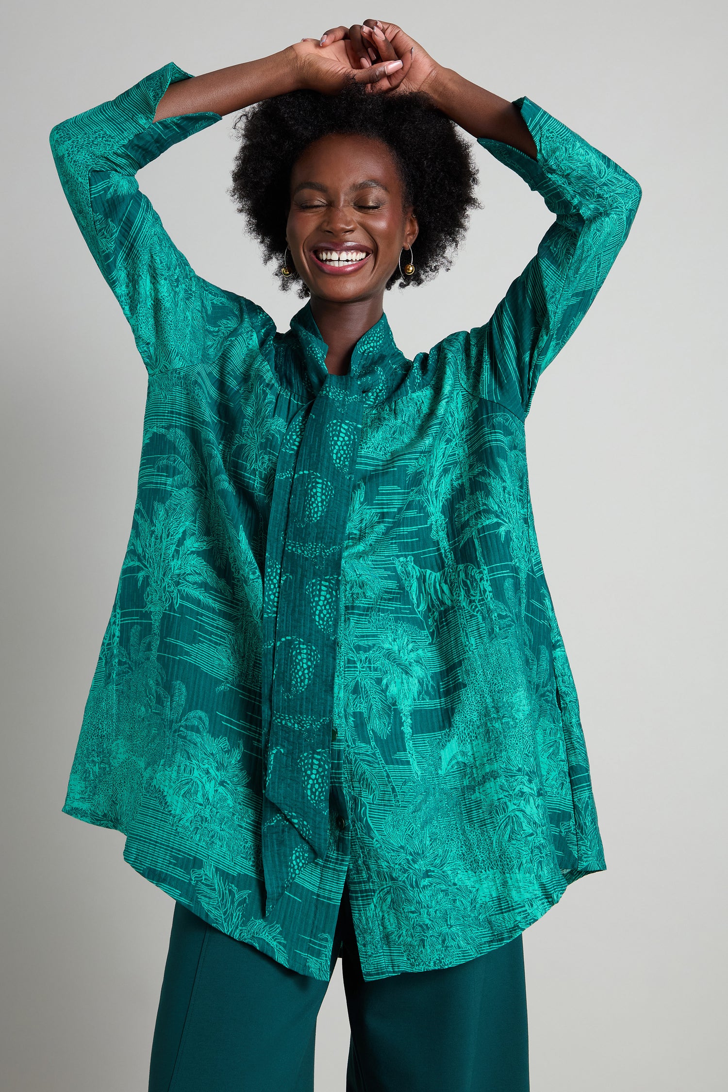 A smiling person dressed in a vibrant Tie Neck Viscose Shirt with arms raised above their head, striking a joyful pose against a plain background.