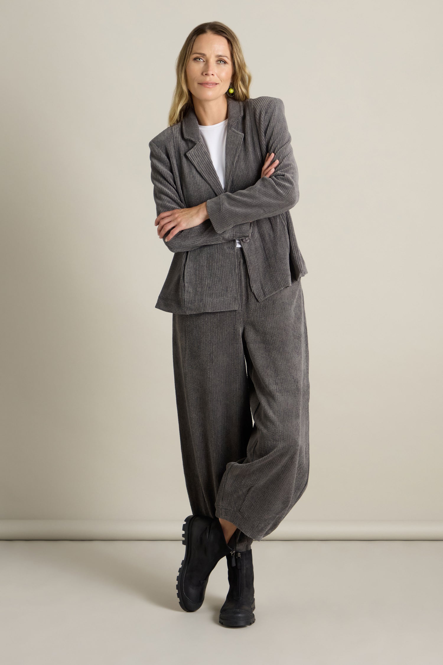 A woman stands confidently with arms crossed, wearing a flattering fit grey corduroy suit and black boots, against a plain background. Her Stretch Cord Fitted Jacket from our latest collection adds a sleek touch to the ensemble.