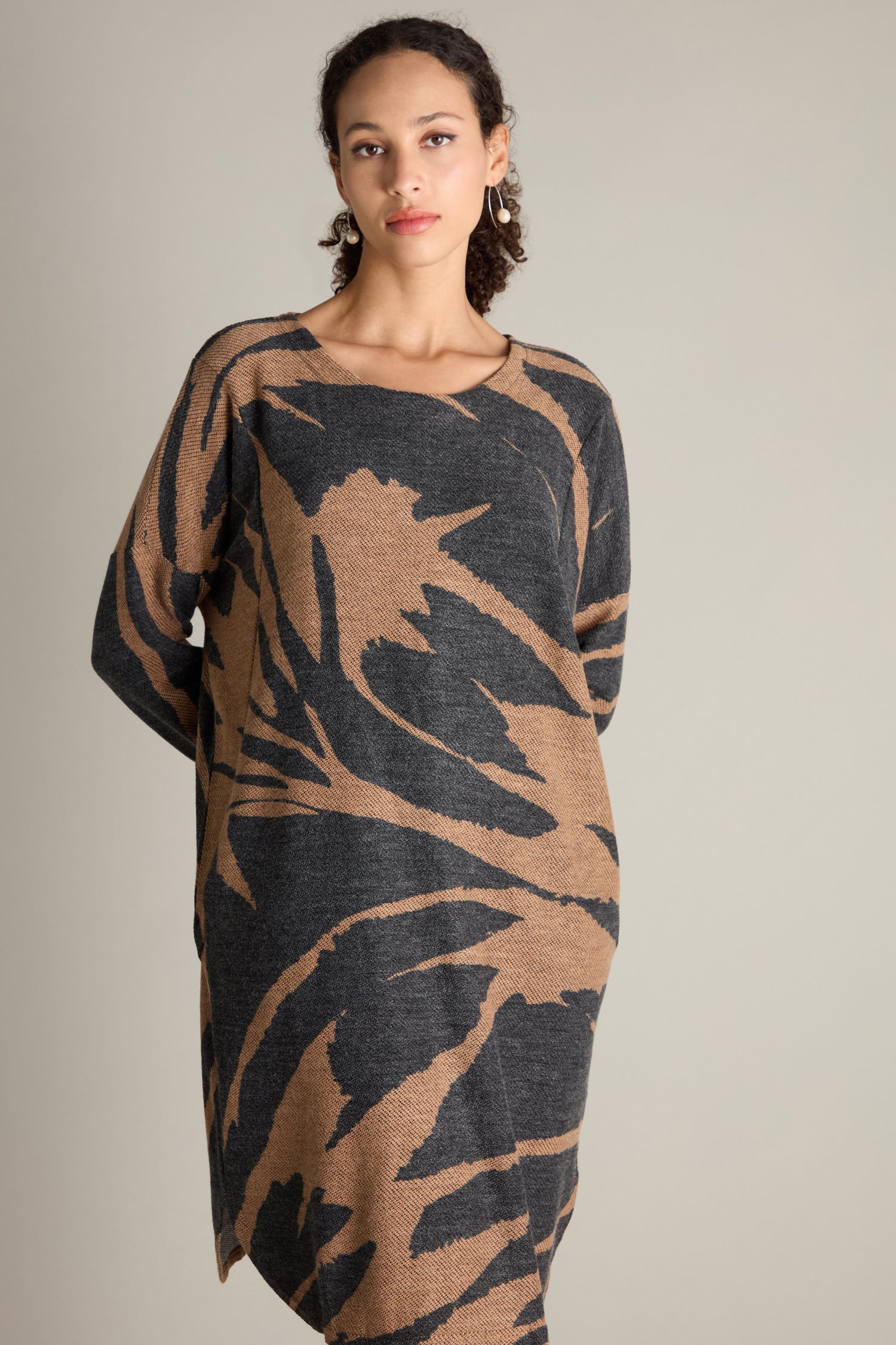 A woman stands against a plain background wearing the Scoop Neck Jacquard Dress, which features long sleeves and an oversized fit with a black and tan abstract pattern. Her expression is neutral, and her hair is tied back.