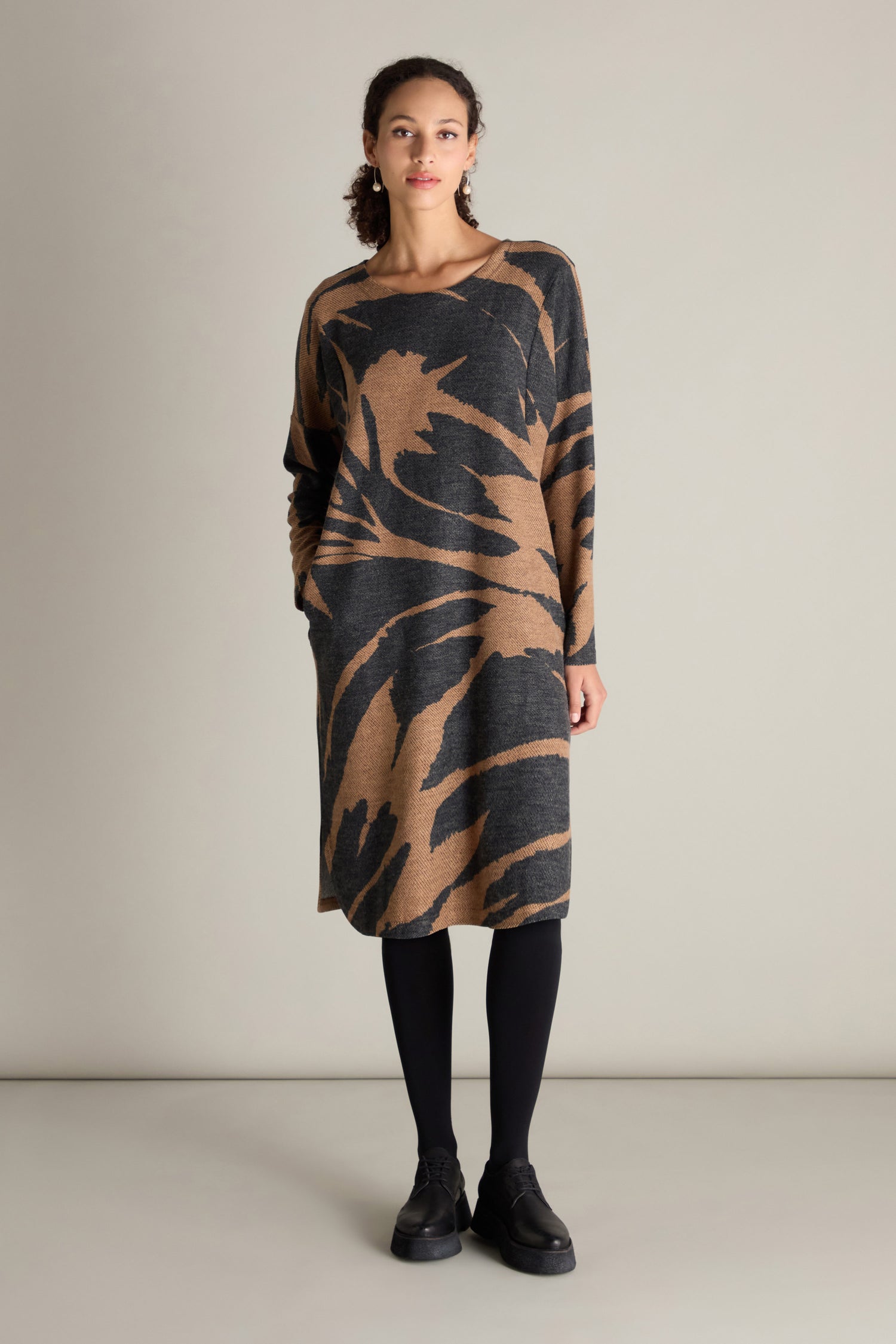 A person stands against a neutral background, dressed in a luxurious Scoop Neck Jacquard Dress featuring intricate brown and black patterns. They have paired the dress with black leggings and shoes, their arms relaxed at their sides.