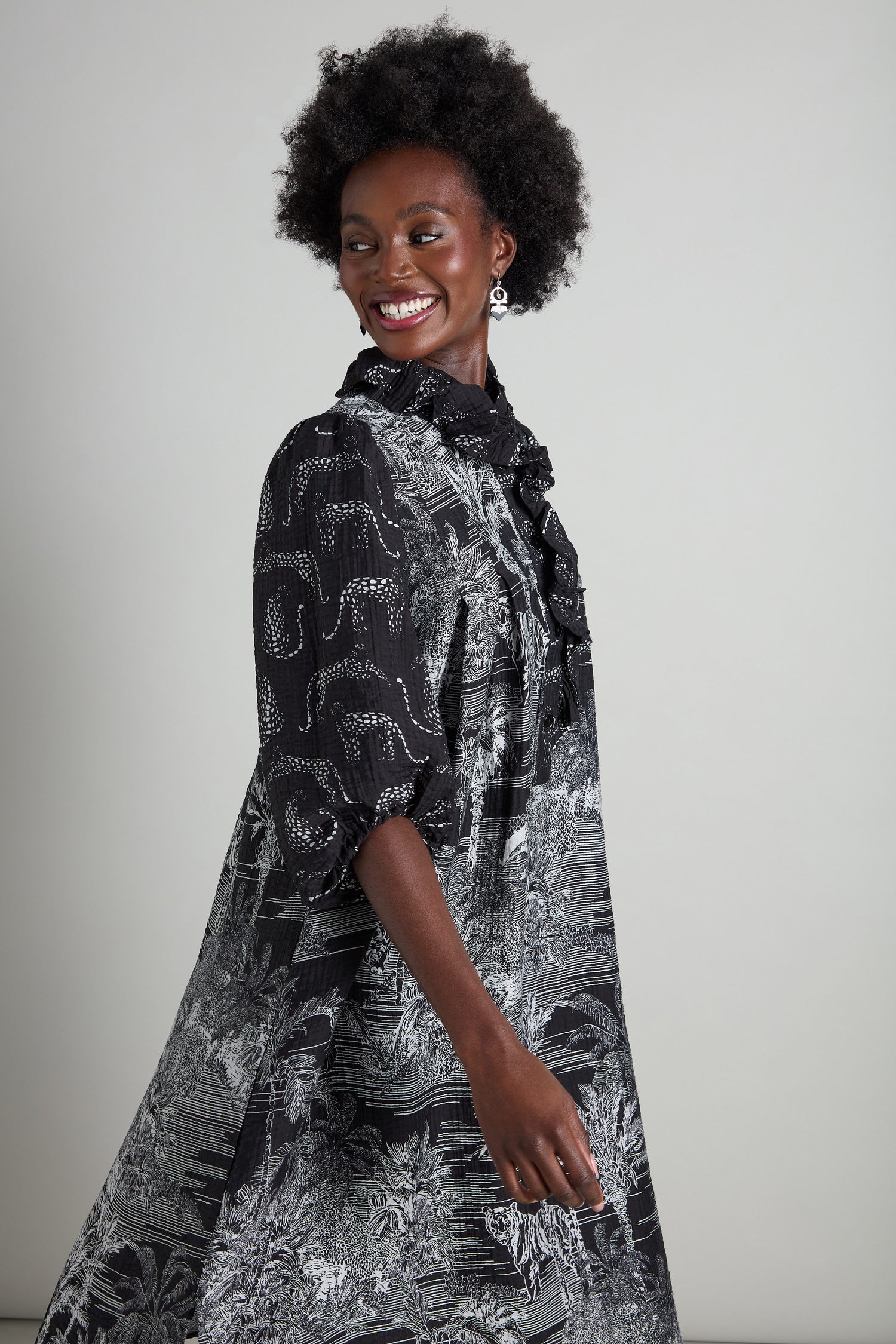 A woman in a Frill Neck Viscose Dress adorned with a jungle-inspired print smiles while looking to the side, highlighting the elegant A-line silhouette.