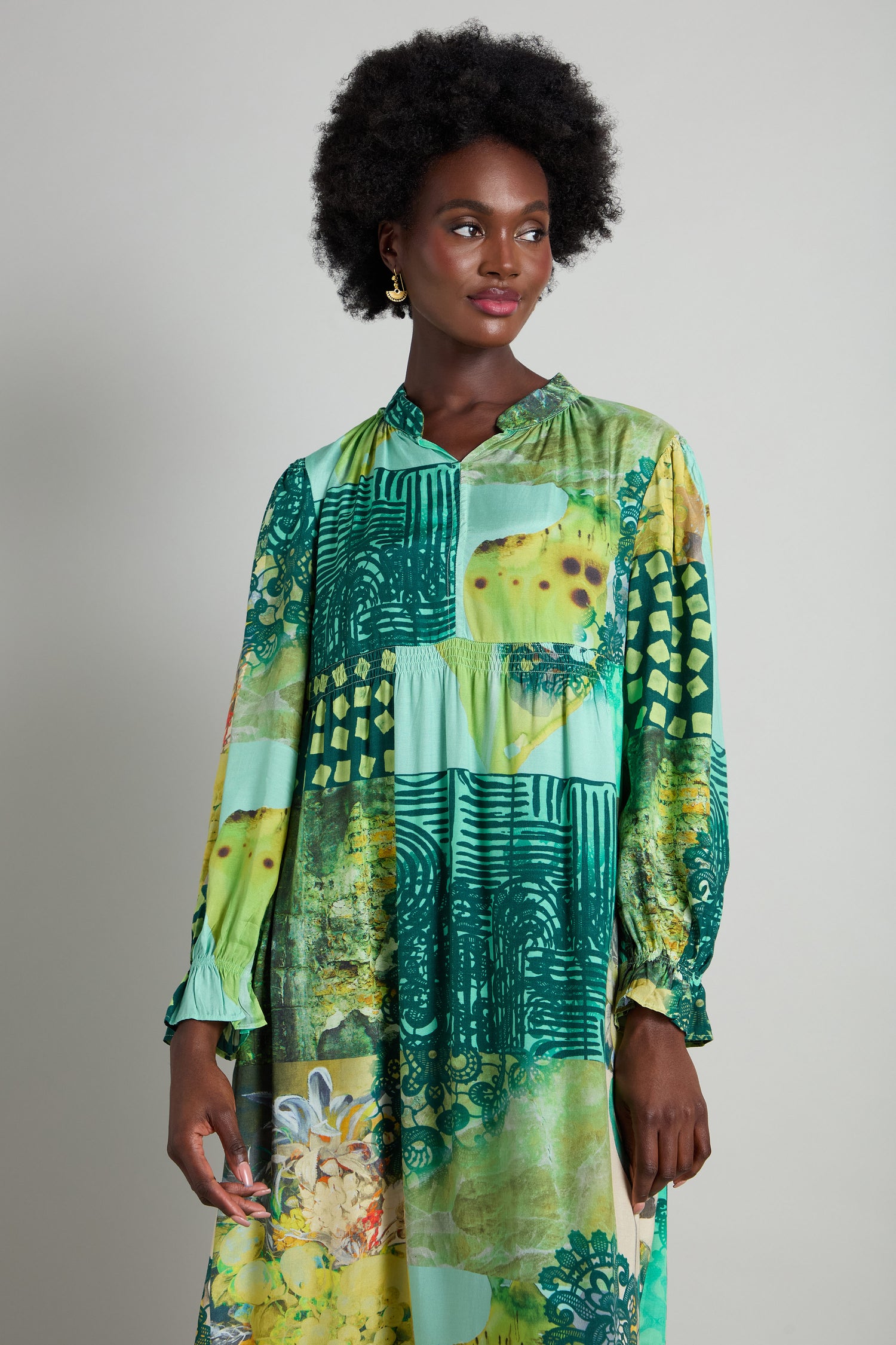 A woman poses against a simple backdrop in the Patchwork Print Viscose Dress. This elegant green dress, featuring long sleeves and an empire waistline, enhances its sophisticated pattern.