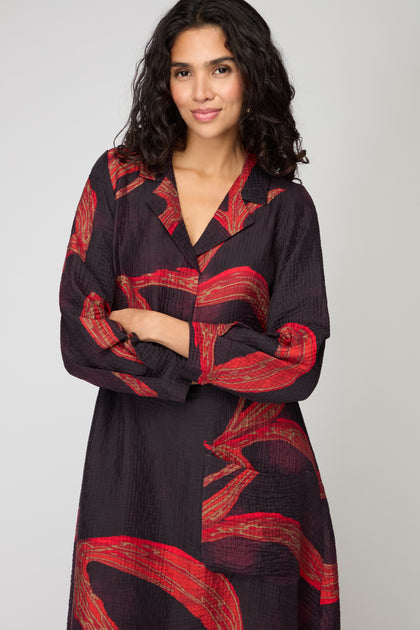 Woman with dark wavy hair wearing a Ribbon Print Viscose Dress, featuring bold red abstract patterns in a Scandinavian A-line midi style, standing with arms crossed and smiling slightly.