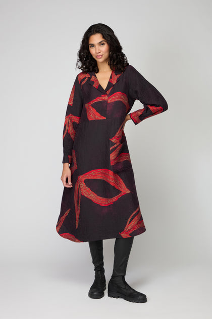 A woman stands against a plain background wearing the Ribbon Print Viscose Dress, a black Scandinavian A-line midi dress with bold red abstract patterns, paired with black leggings and boots.