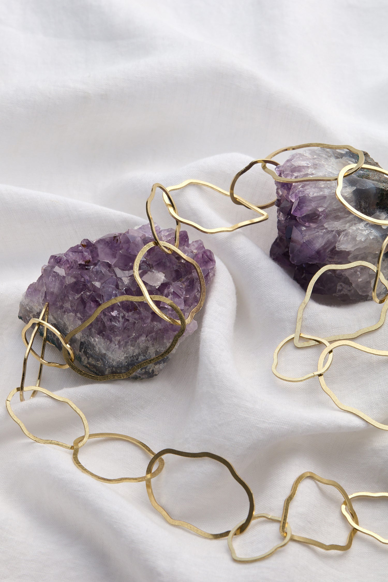 The Crushed Hoop Necklace, a lightweight gold piece adorned with two stunning purple amethyst stones, gently rested on a pristine white fabric.