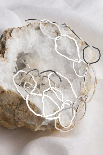 The Crushed Hoop Necklace features abstract metal wire art in gold or silver, elegantly displayed on a piece of quartz crystal and set against a white fabric background, creating a lightweight and sophisticated accessory.