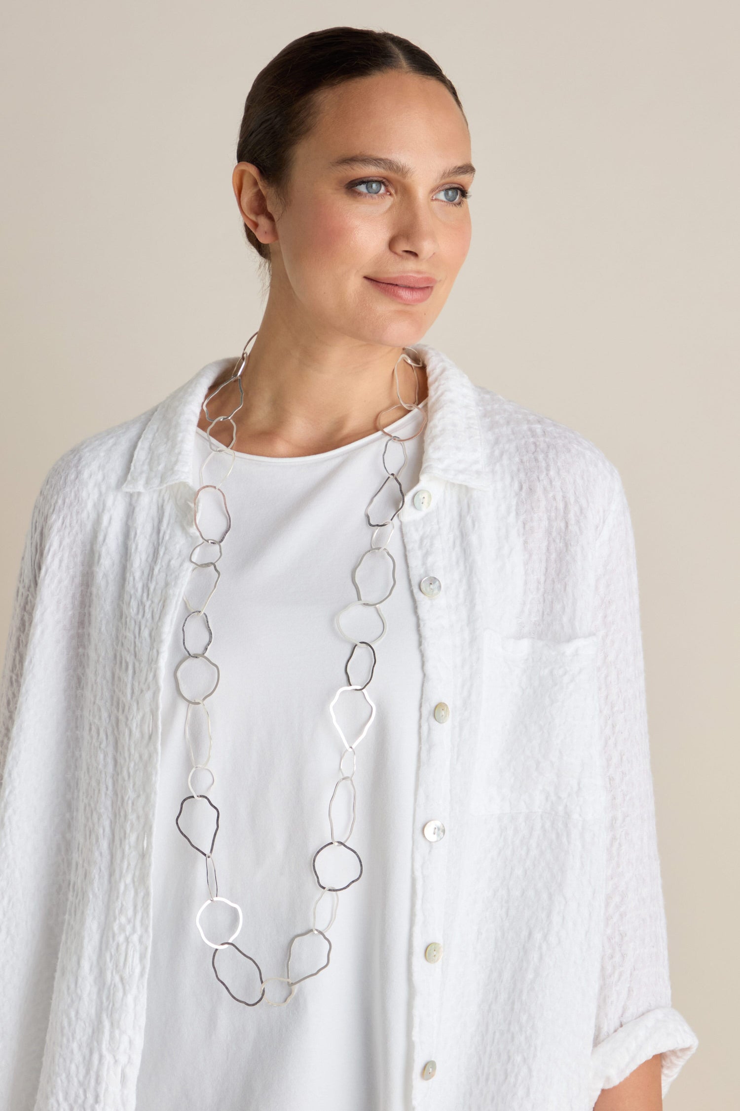 A woman wearing a white textured shirt and a long Crushed Hoop Necklace, featuring contemporary geometric shapes, looks to her left against a plain background.