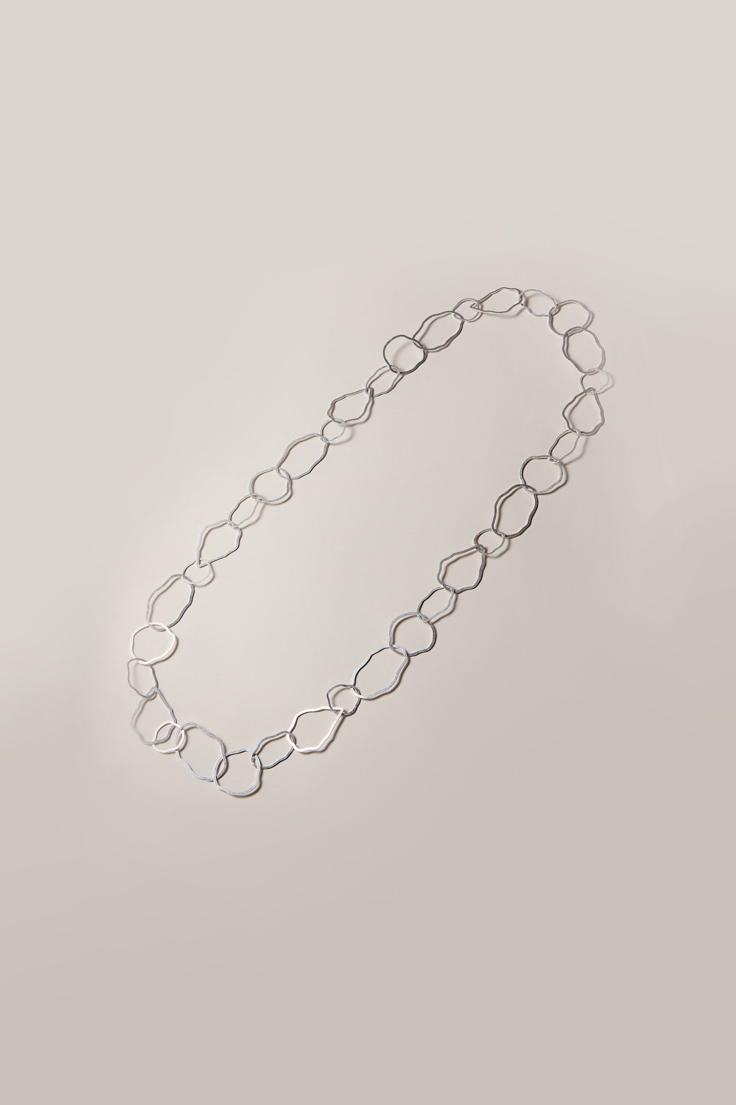 The Crushed Hoop Necklace is a lightweight silver chain featuring interlocking geometric-shaped links against a plain background.