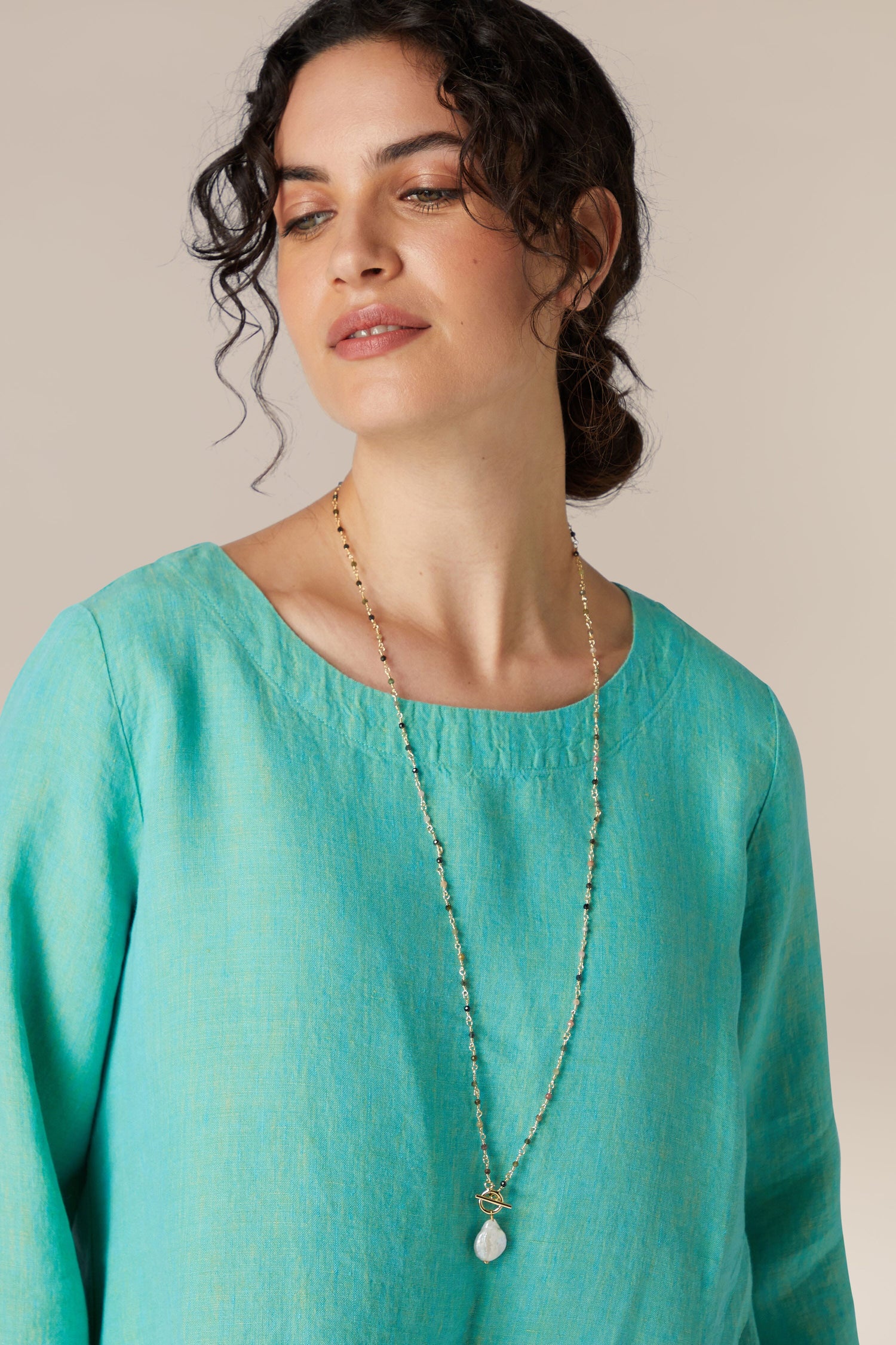 Sentence with product name: Woman with curly hair wearing a turquoise blouse and a delicate necklace featuring a Cultured Pearl Pendant, adding an elegant touch.