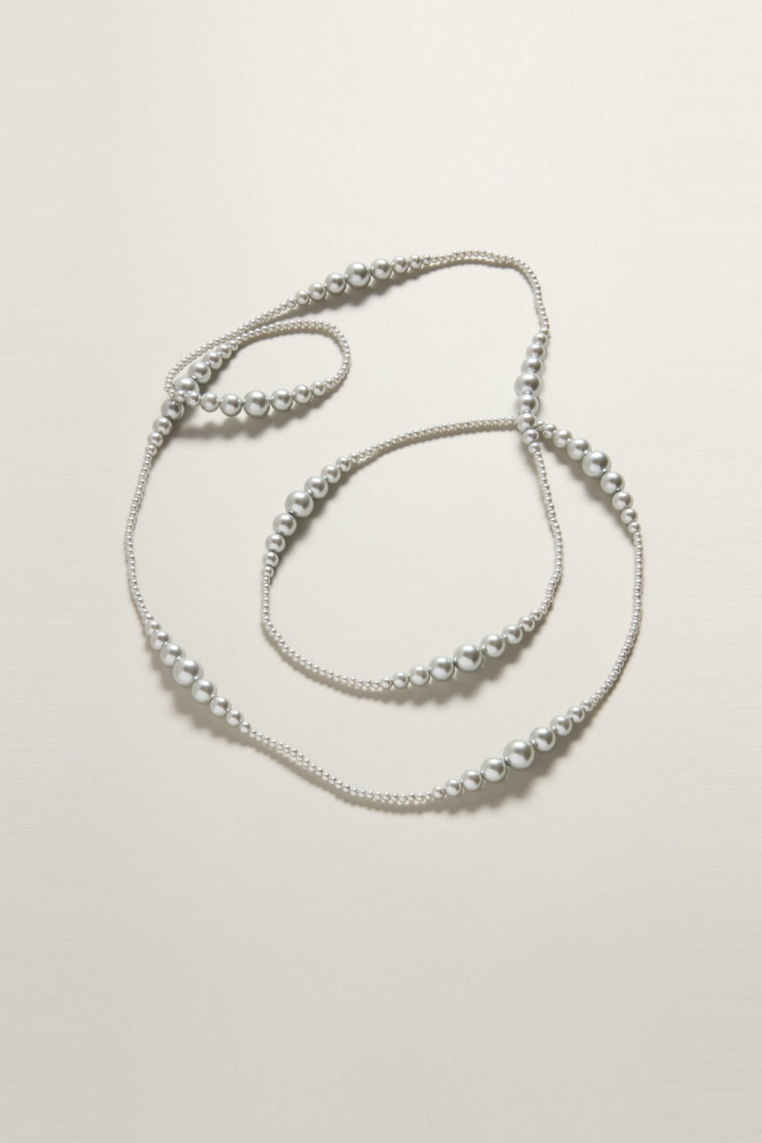 The Graduated Pearl Necklace features a long, silver strand with beads of varying sizes, displaying timeless elegance against a plain, light background.