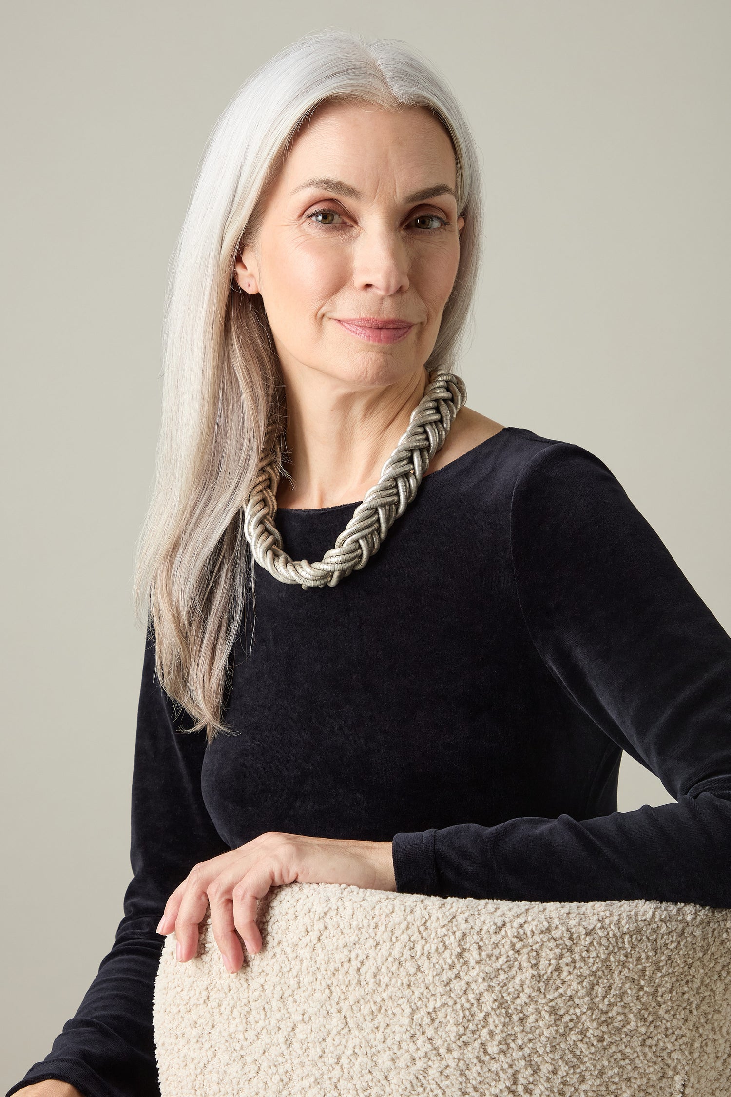 An older woman with long gray hair sits gracefully on a textured chair, embodying contemporary elegance in a black top accented by the Silver Braided Necklace. Her statement piece showcases artisanal craftsmanship as she gazes confidently at the camera.