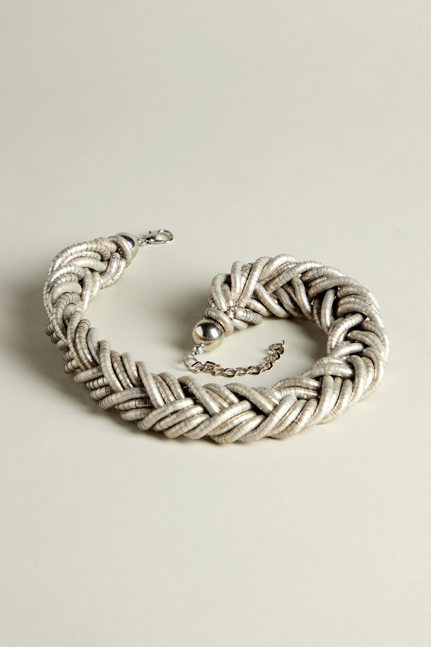 The Silver Braided Necklace rests elegantly on a light gray surface, showcasing its contemporary allure with a woven metal design, complete with a clasp and a short dangling chain.