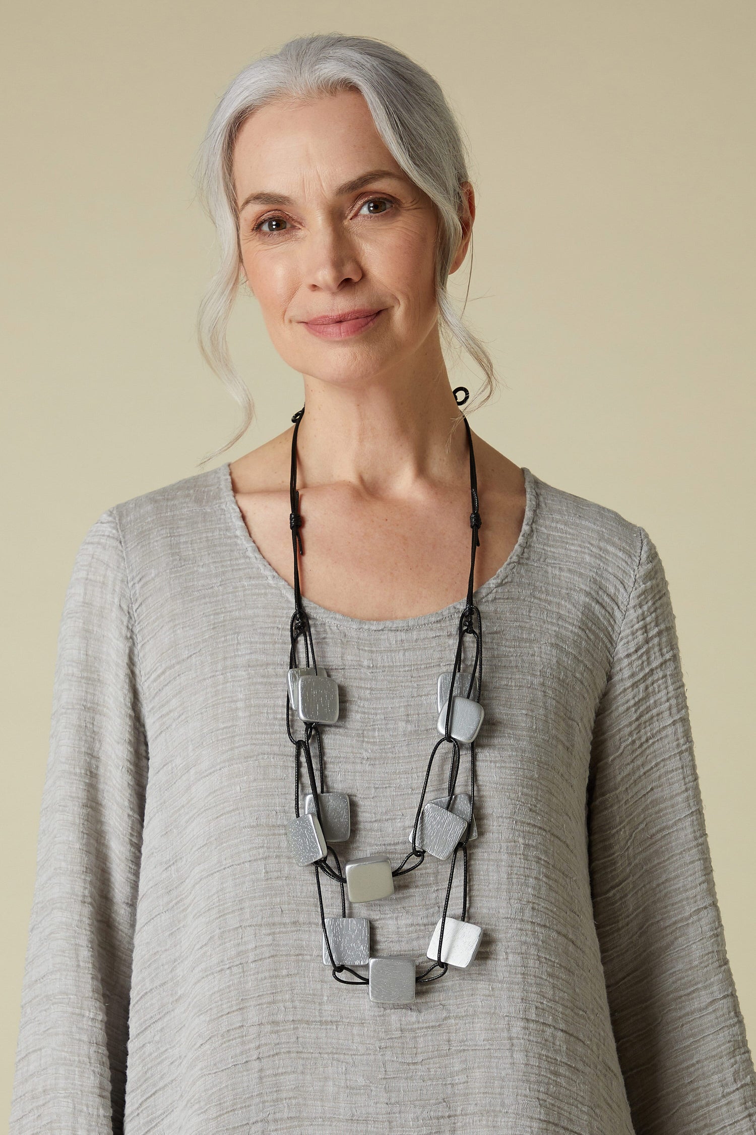 A mature woman with gray hair, wearing a gray top and an impactful Classic Chain Necklace in brushed silver, smiling gently at the camera.