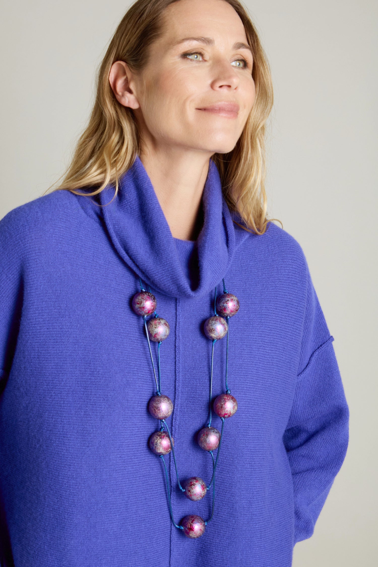 A woman, adorned in a purple sweater and the striking Metallic Planets Necklace featuring large, colorful beads, gazes slightly upward with a soft smile, emanating an air of cosmic elegance.