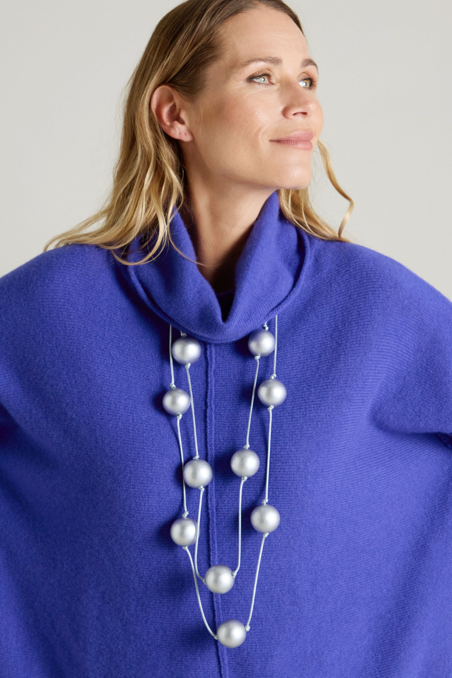 A person is shown wearing a blue high-neck garment and a Metallic Planets Necklace adorned with large silver spheres, exuding cosmic elegance as they gaze off to the side.