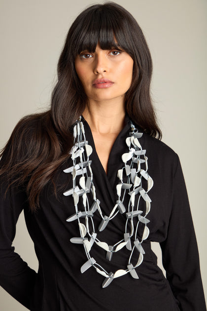 A person with long hair is wearing the Metallic Shapes Necklace, which features layered, geometric silver elements combined with a soft cord, over a black top against a neutral background.