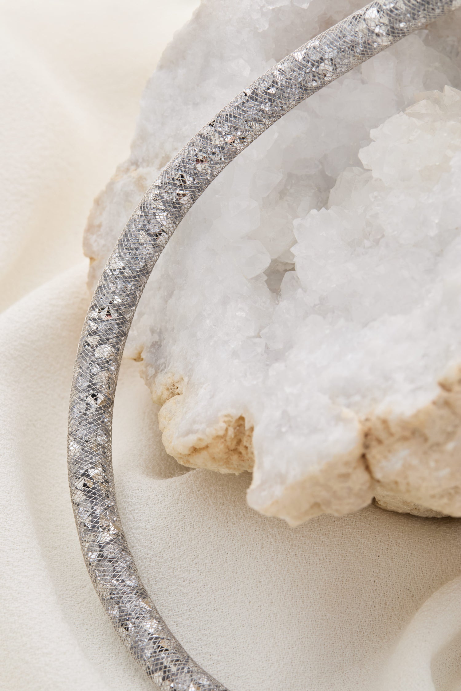 Close-up of the Short Diamante Necklace, featuring a textured silver design adorned with shimmering diamante stones, resting on a white crystalline mineral formation and set against a beige fabric background.