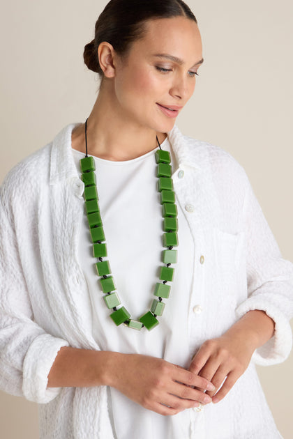 Person wearing a white outfit and the Long Cubes Necklace with green resin cubes, looking down and smiling softly against a plain background.