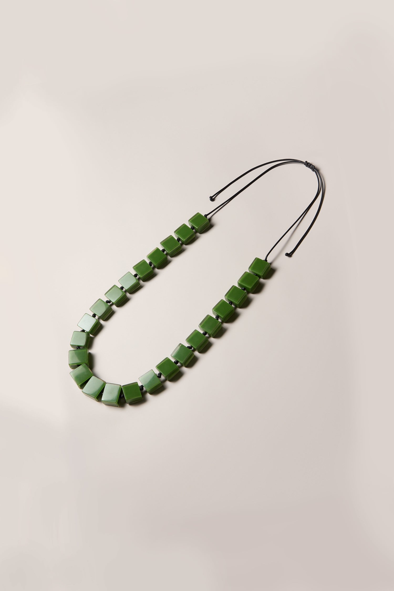 The Long Cubes Necklace, with its green square beads and black string, arranged on a beige background, is perfect for a daytime look.