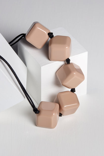 The Twisted Cube Cord Necklace, a unique piece featuring an adjustable black cord adorned with five beige, cube-shaped beads, is elegantly displayed on white geometric blocks.