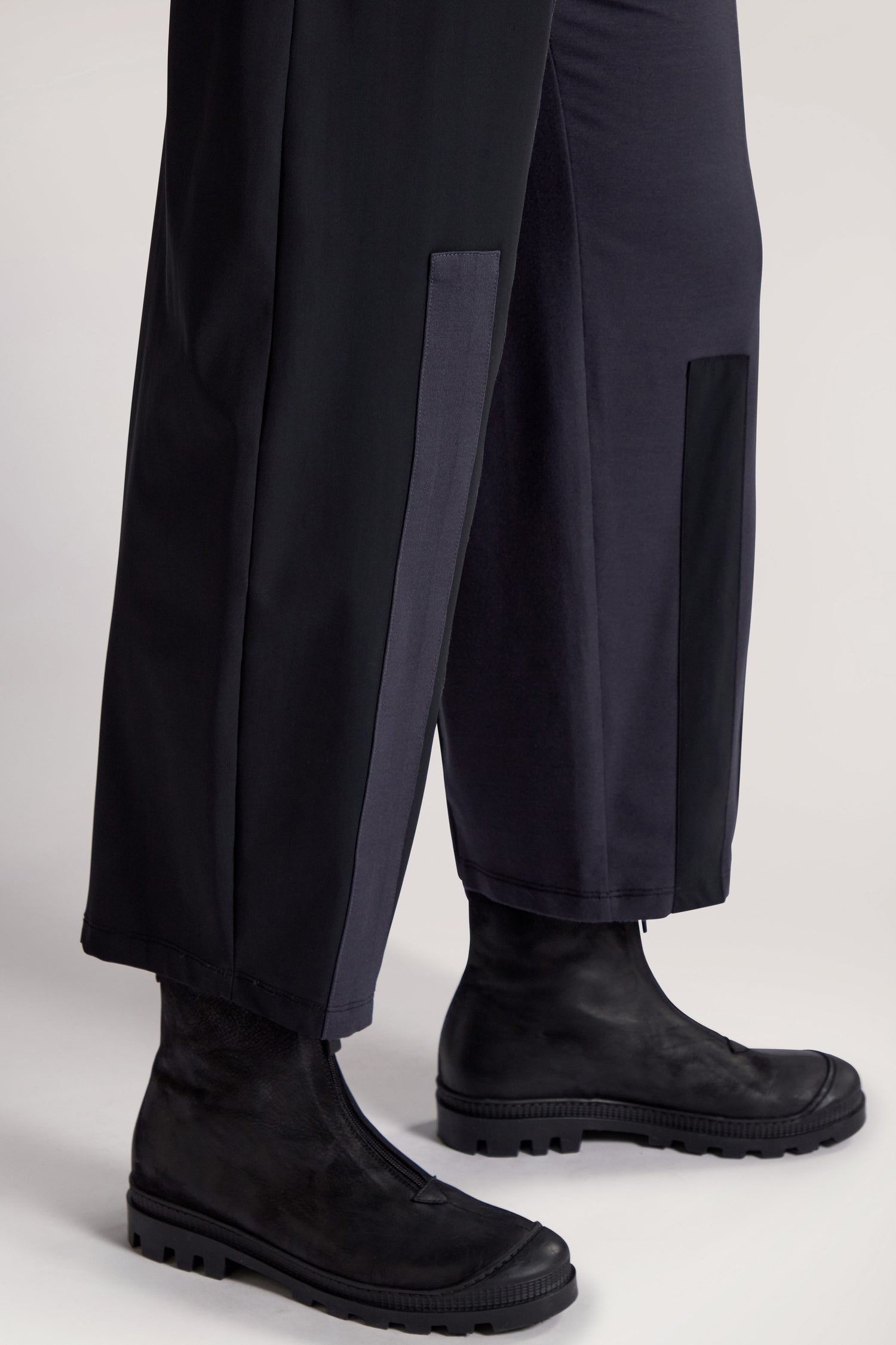 A person wearing Panelled Jersey Trousers and black ankle boots stands on a light-colored surface. The relaxed wide-leg silhouette of the trousers is adorned with rectangular patches at the bottom.