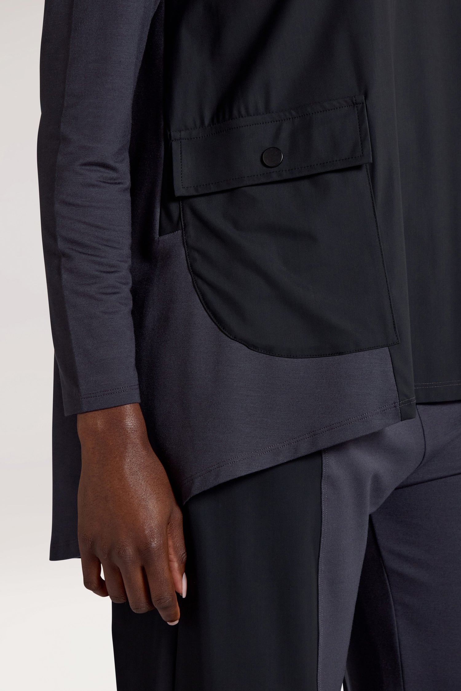 Close-up of a person wearing a black Zip Collar Pocket Top, featuring a large flap pocket on the side of their shirt. Only the lower half of their body and one arm are visible, showcasing the sleek design and functionality.