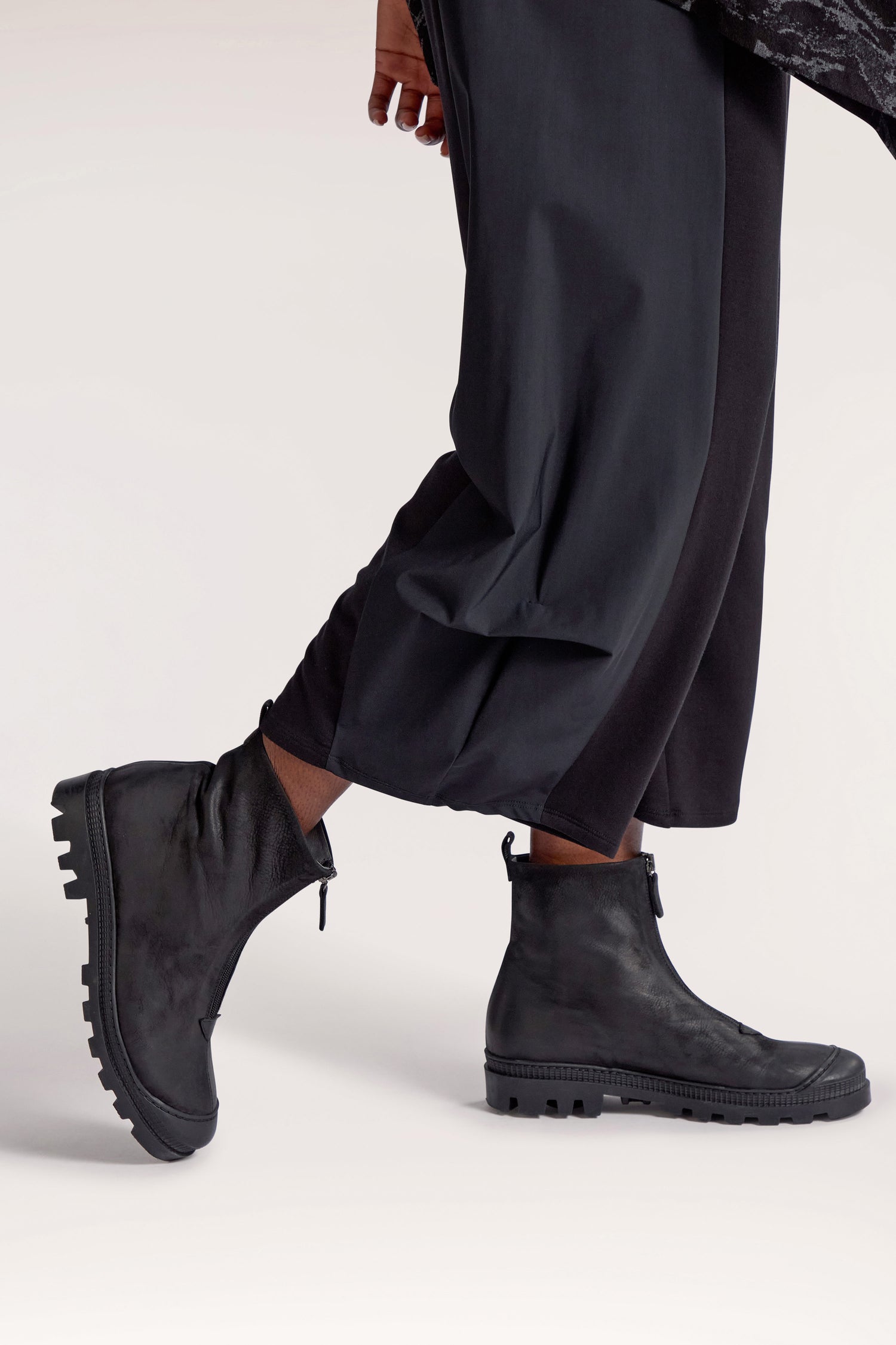 A person wearing Side Panel Pleat Hem Trousers with an elasticated waistband and black, high-ankle boots with rugged soles stands against a plain background.