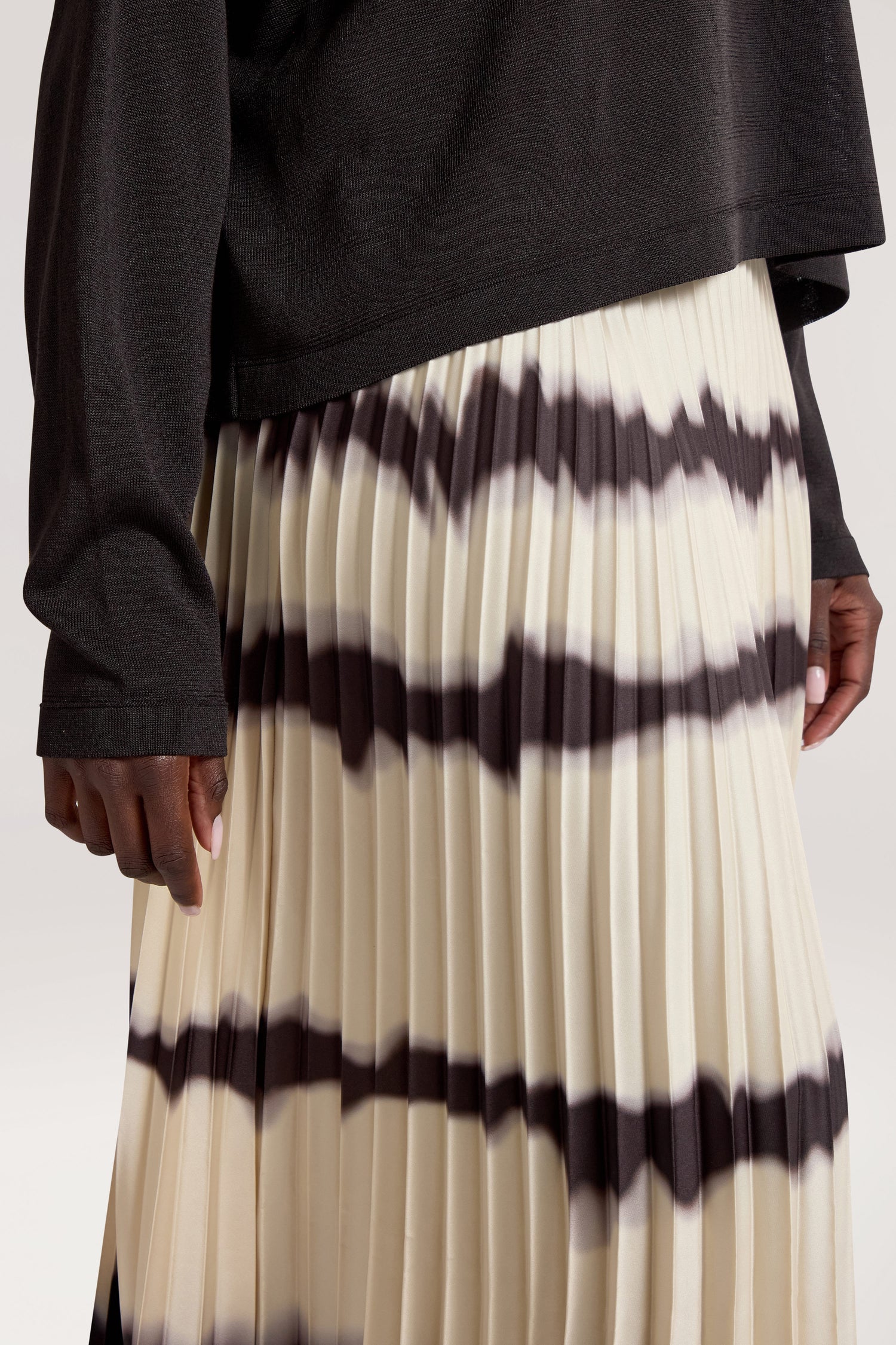 A person is shown wearing a Tie Dye Pleat Dress With Jersey Top, featuring a black long sleeve jersey top and a pleated skirt with horizontal black and white stripes. The image focuses on the lower torso and the upper part of the skirt.