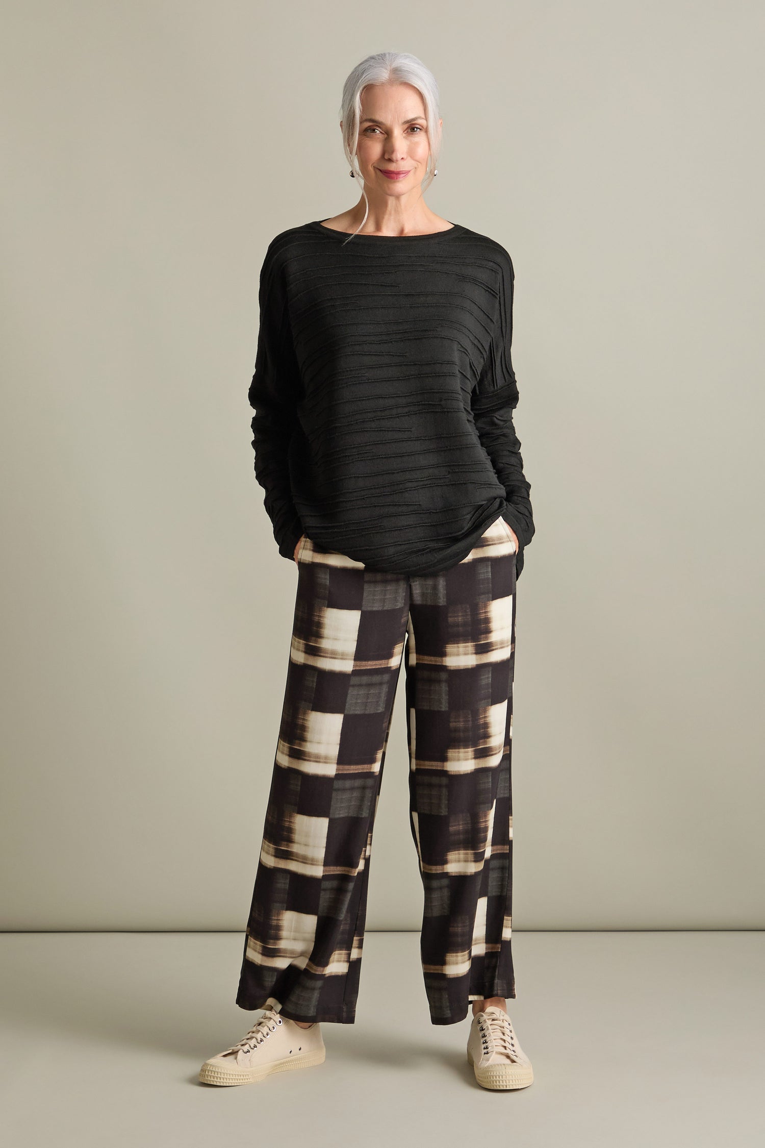 A woman with gray hair wears the Rib Detail Wide Neck Knit sweater in black, along with patterned wide-leg pants and beige sneakers, standing with her hands in her pockets against a plain backdrop.