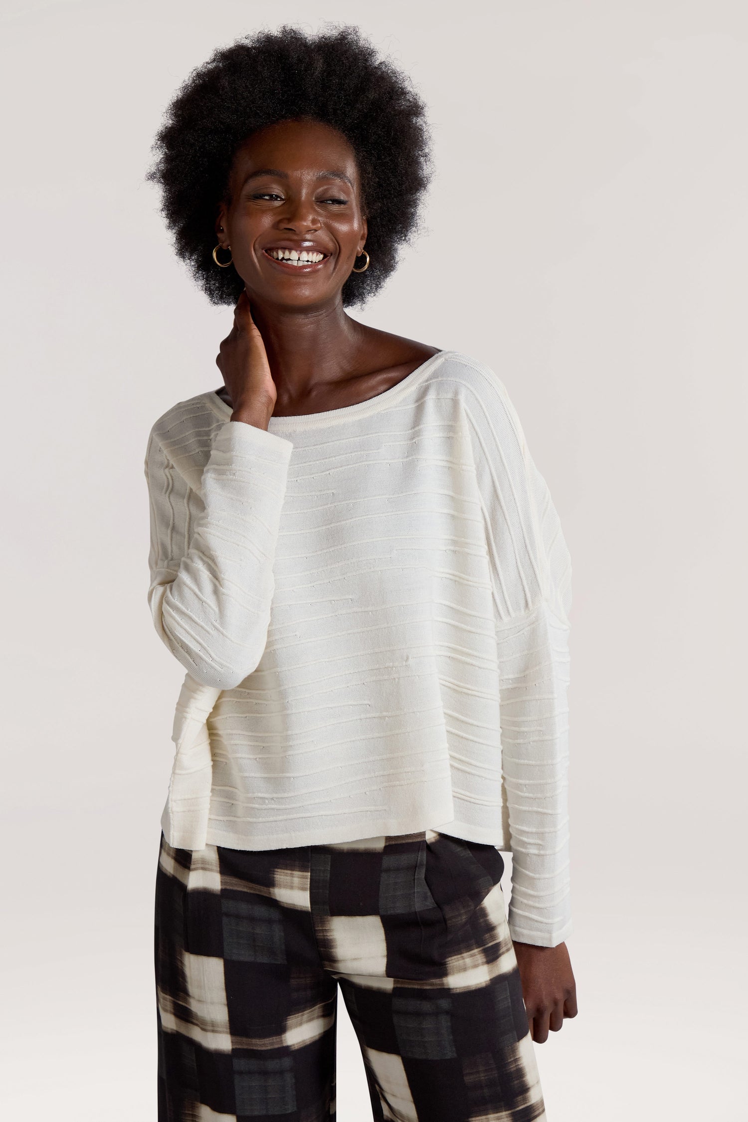 A person with short curly hair smiles while wearing a Rib Detail Wide Neck Knit in white paired with black-and-white plaid pants.