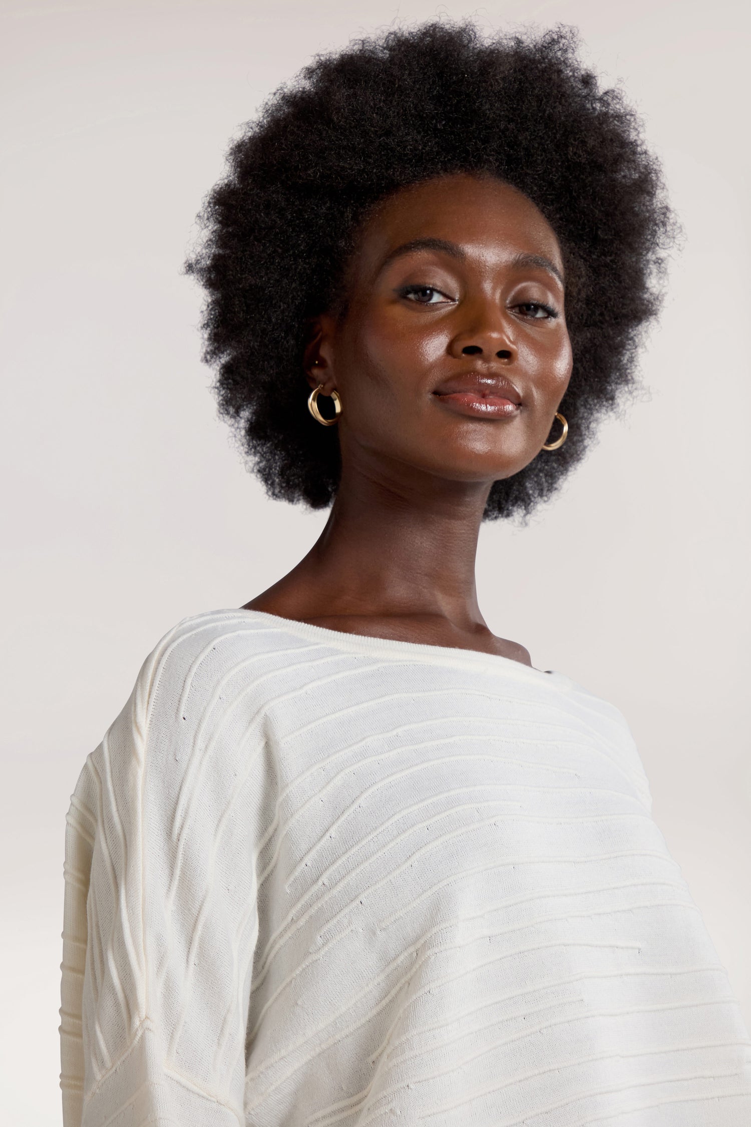 A person with an afro hairstyle, wearing gold hoop earrings and a white Rib Detail Wide Neck Knit top, looks confidently at the camera against a neutral background.