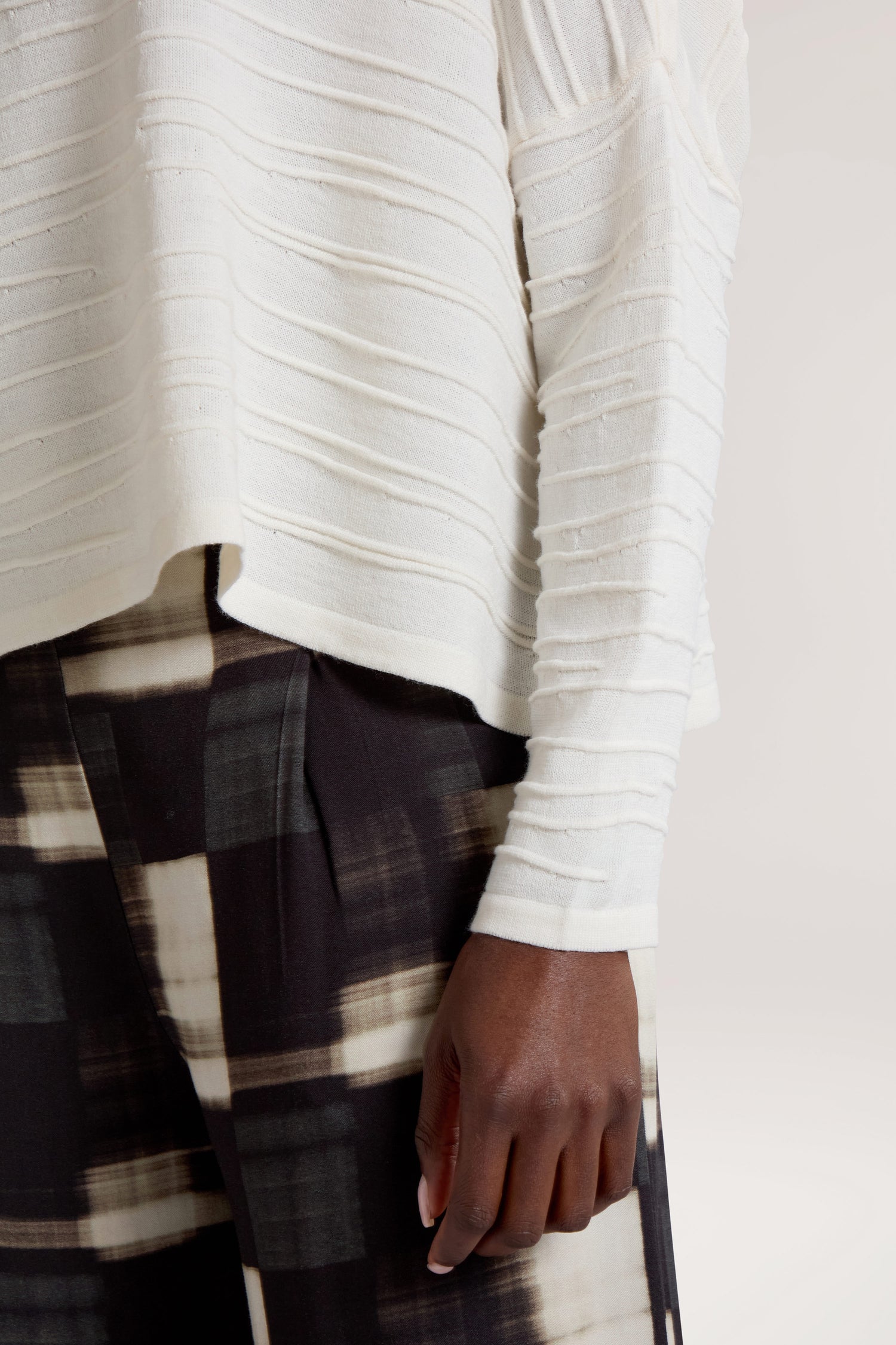 A close-up of a person wearing a cream-colored Rib Detail Wide Neck Knit with textured stripes and black and white plaid pants. The person's right hand, resting against their leg, showcases their minimalist wardrobe.