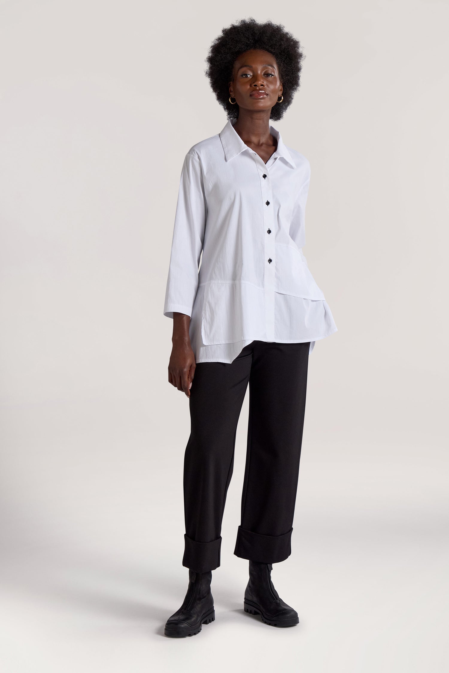 A person with short, curly hair stands facing forward, wearing a white Panel Hem Shirt paired with black pants and black boots against a plain background.